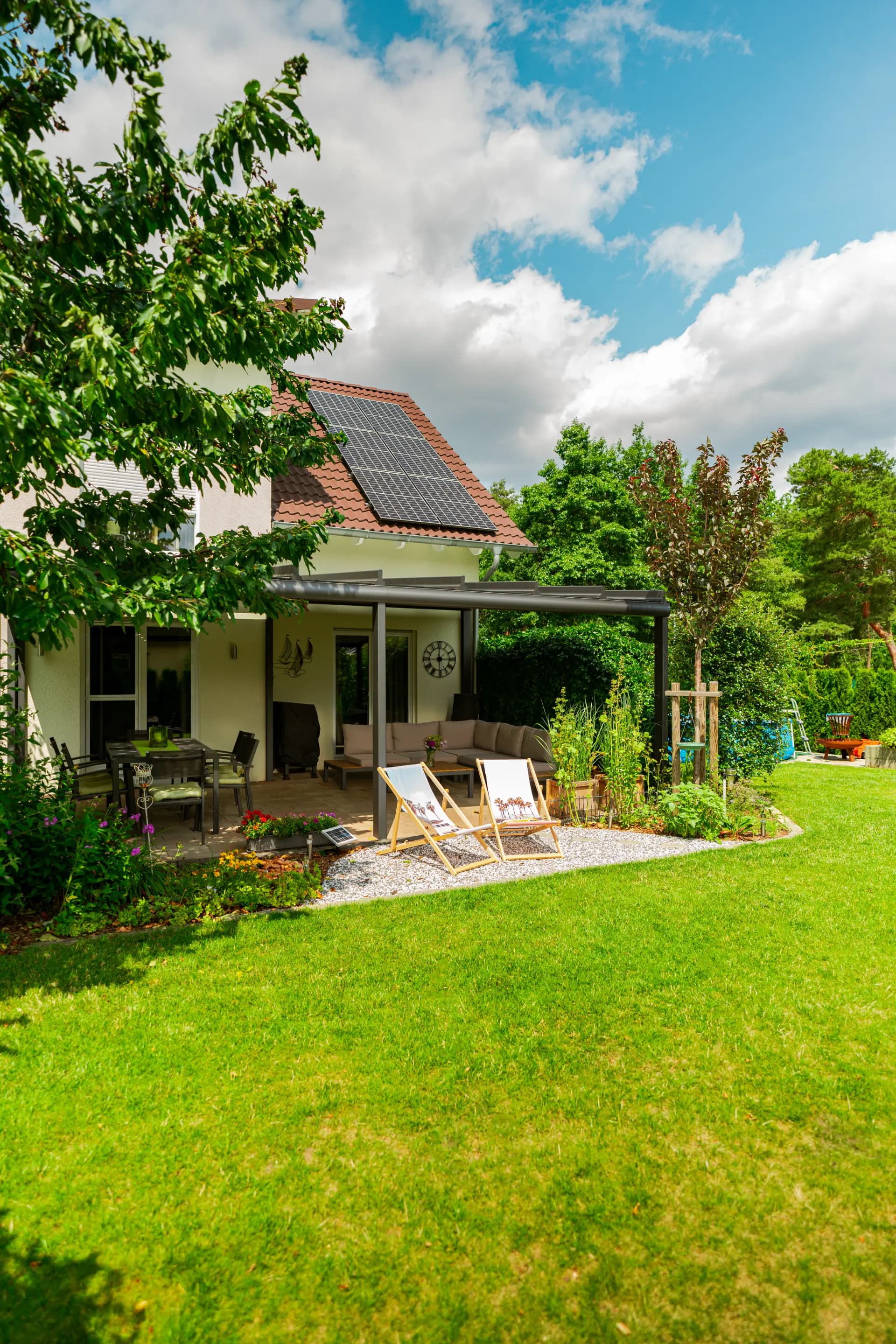 Offenes Terrassendach an einem Einfamilienhaus, darunter eine Sitzgruppe, davor zwei Liegestühle im Garten