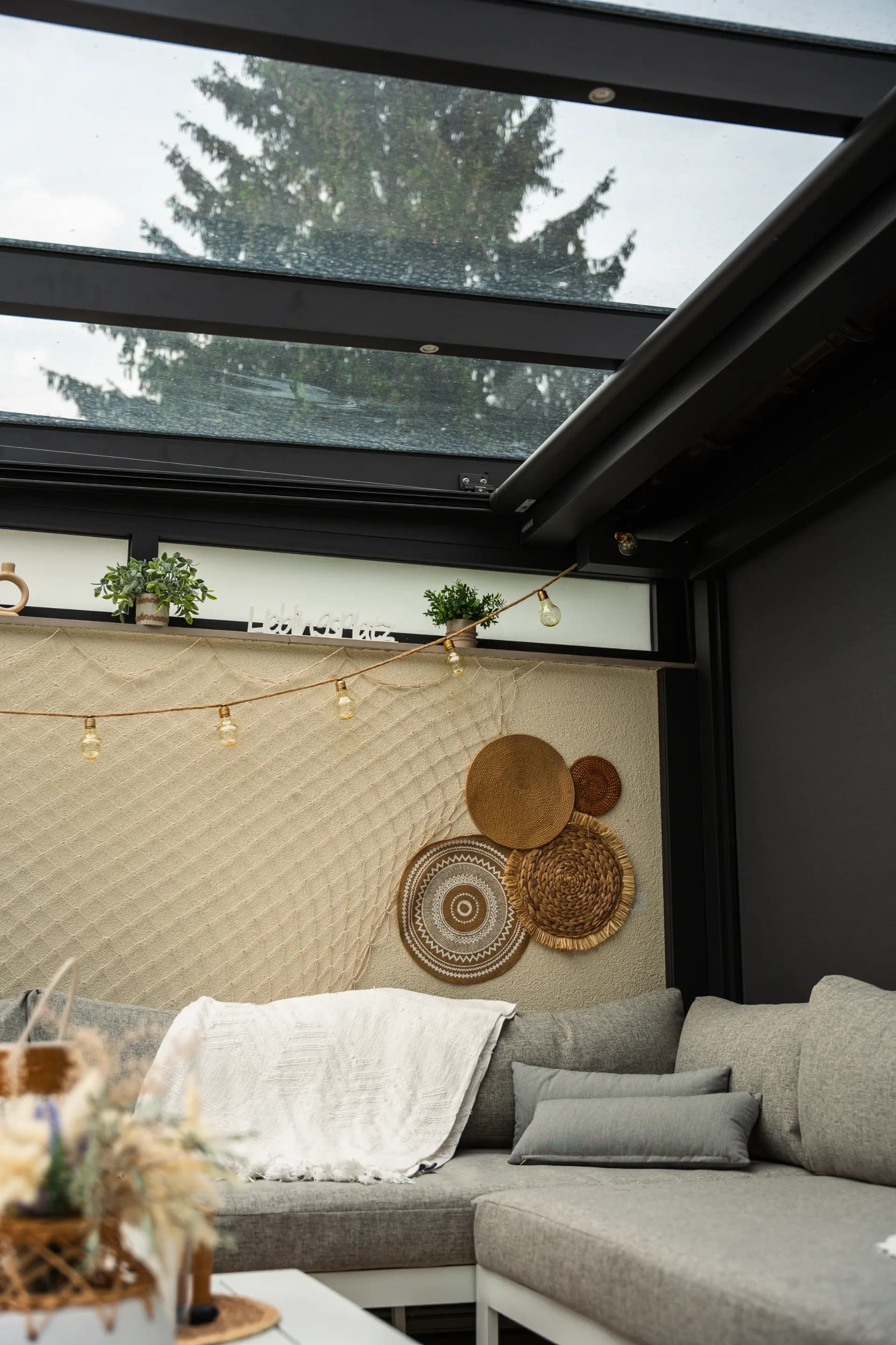 Blick von unten auf ein Glasdach mit Lichterkette über einer Sofaecke mit Kissen und Decke