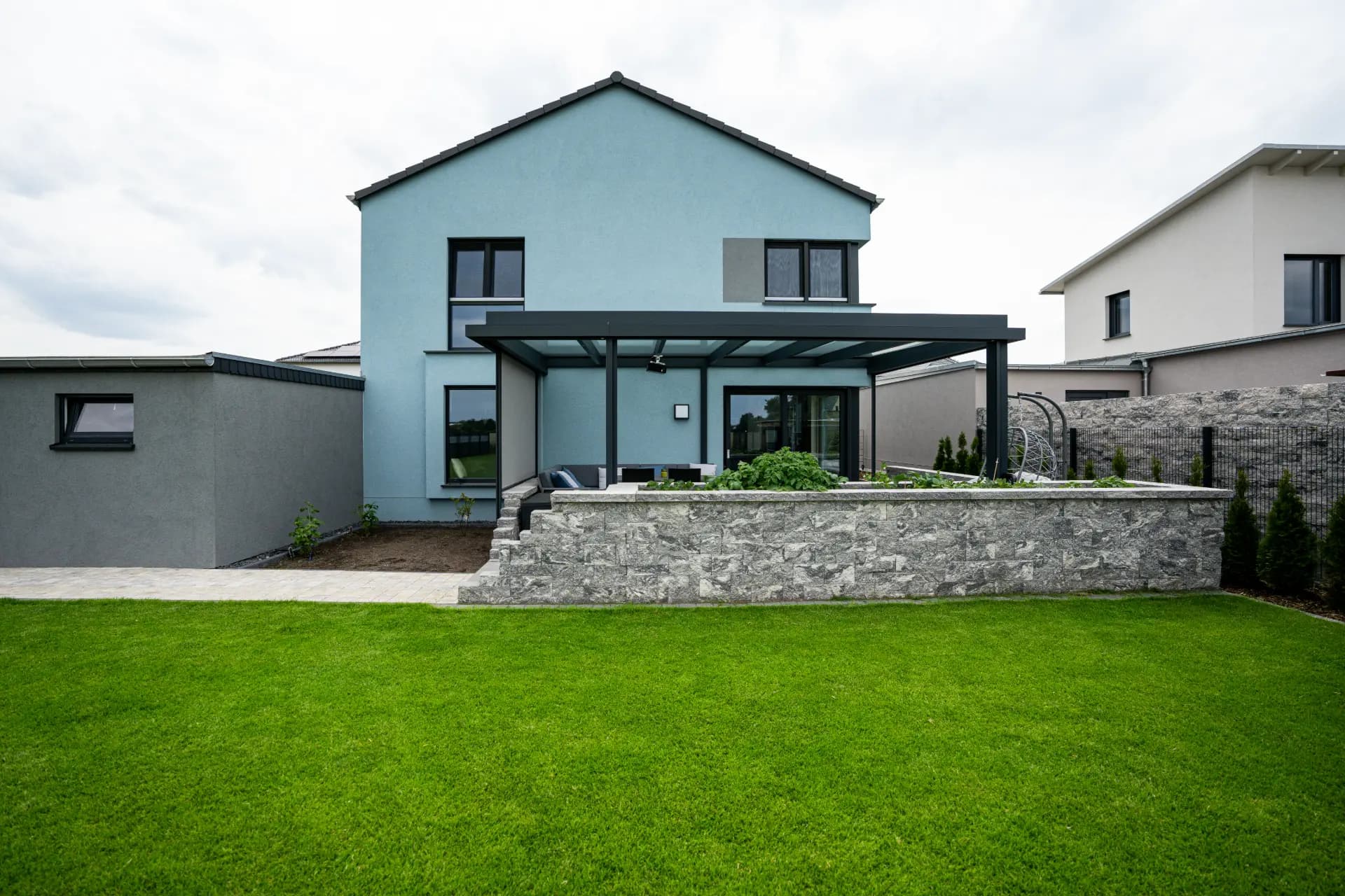Modernes Wohnhaus mit hellblauer Fassade, davor eine überdachte Sitzecke und ein Hochbeet aus Naturstein