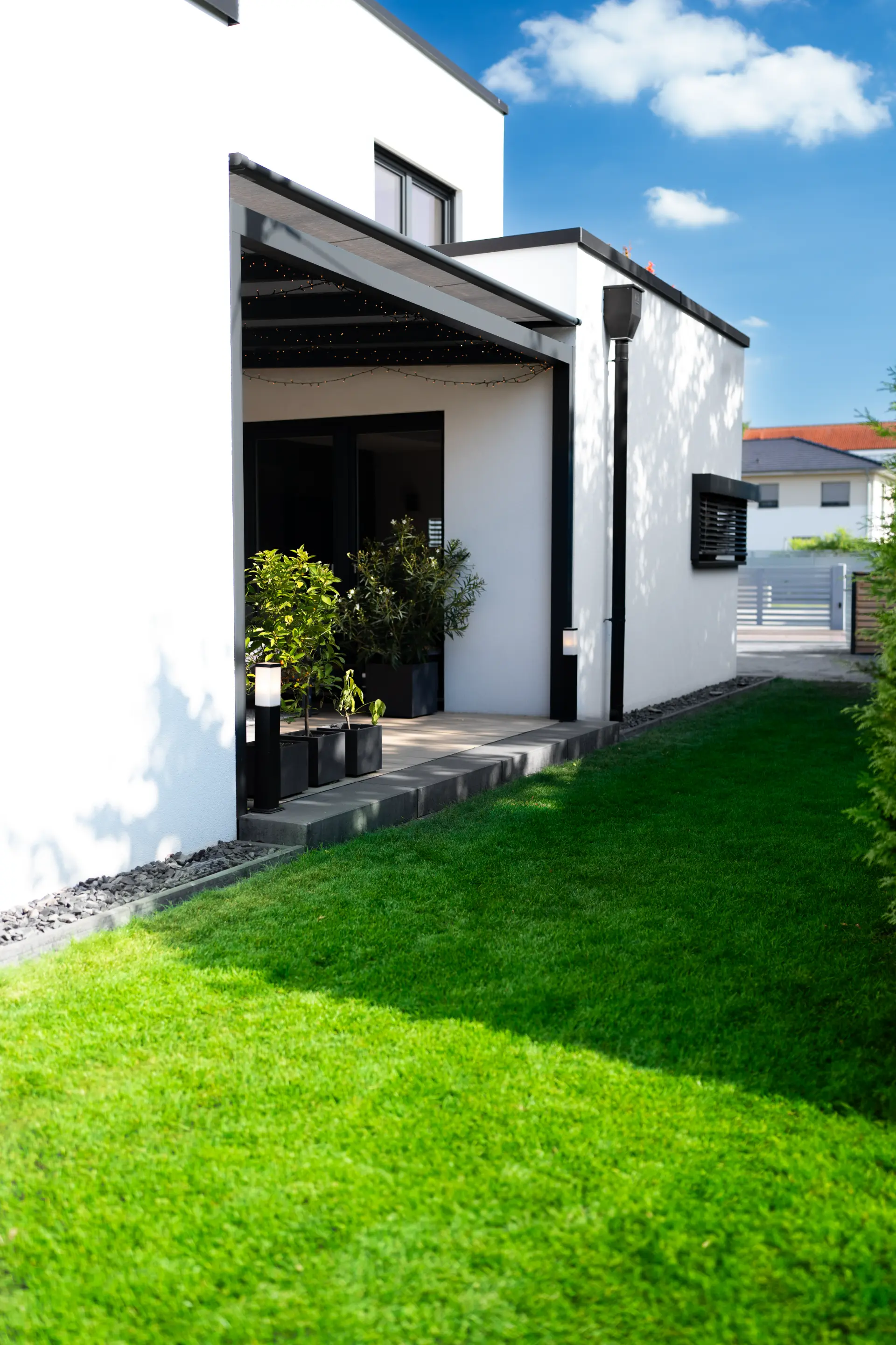 Kleines Terrassendach an moderner weißer Hausecke mit Topfpflanzen und Rasen