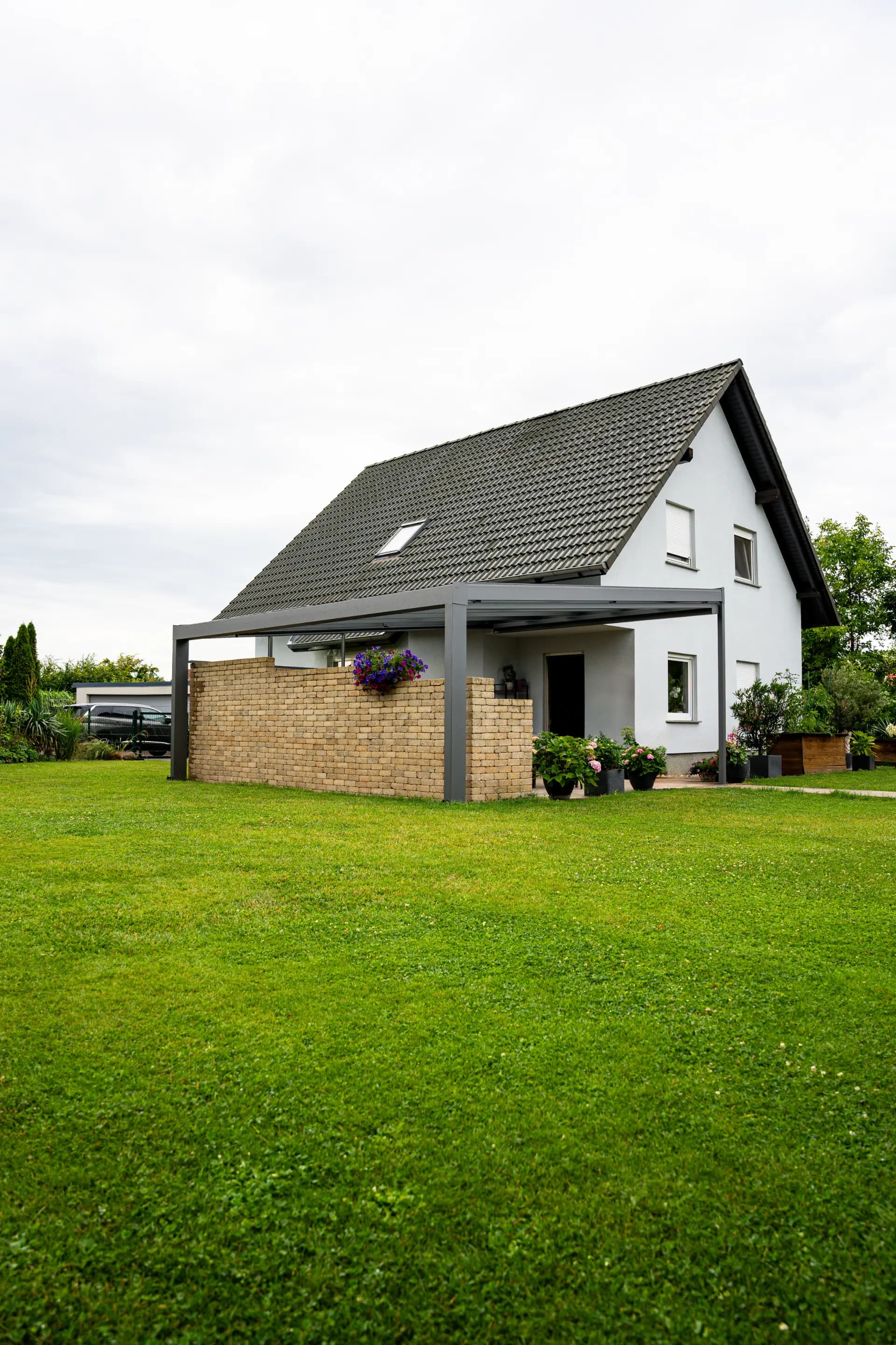 Terrassendach mit Klinker-Sichtschutzwand an einem weißen Einfamilienhaus mit Satteldach, davor blühende Petunien