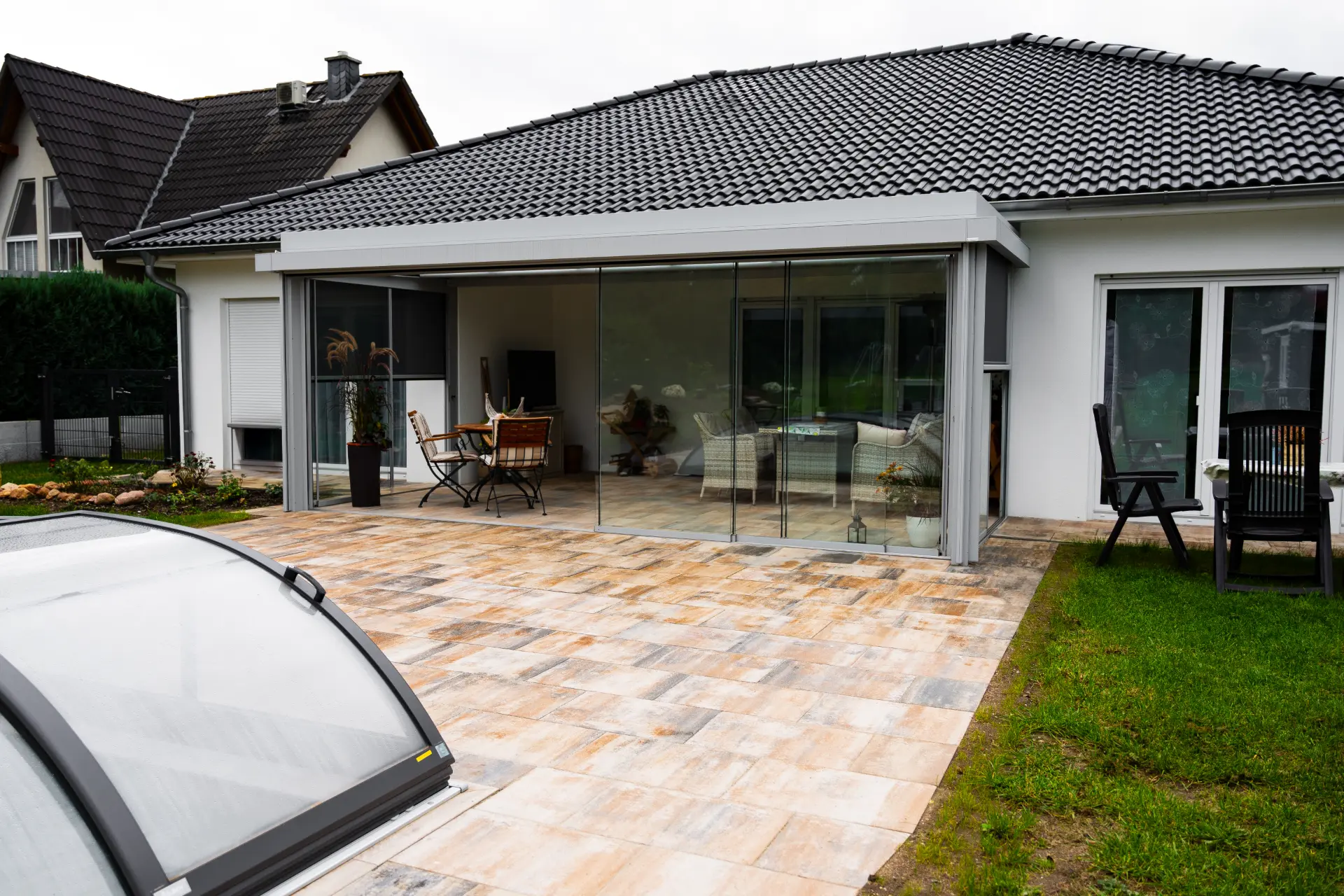 Terrassendach mit geschlossenen Glasschiebewänden an einem weißen Bungalow, dahinter das möblierte Wohnzimmer