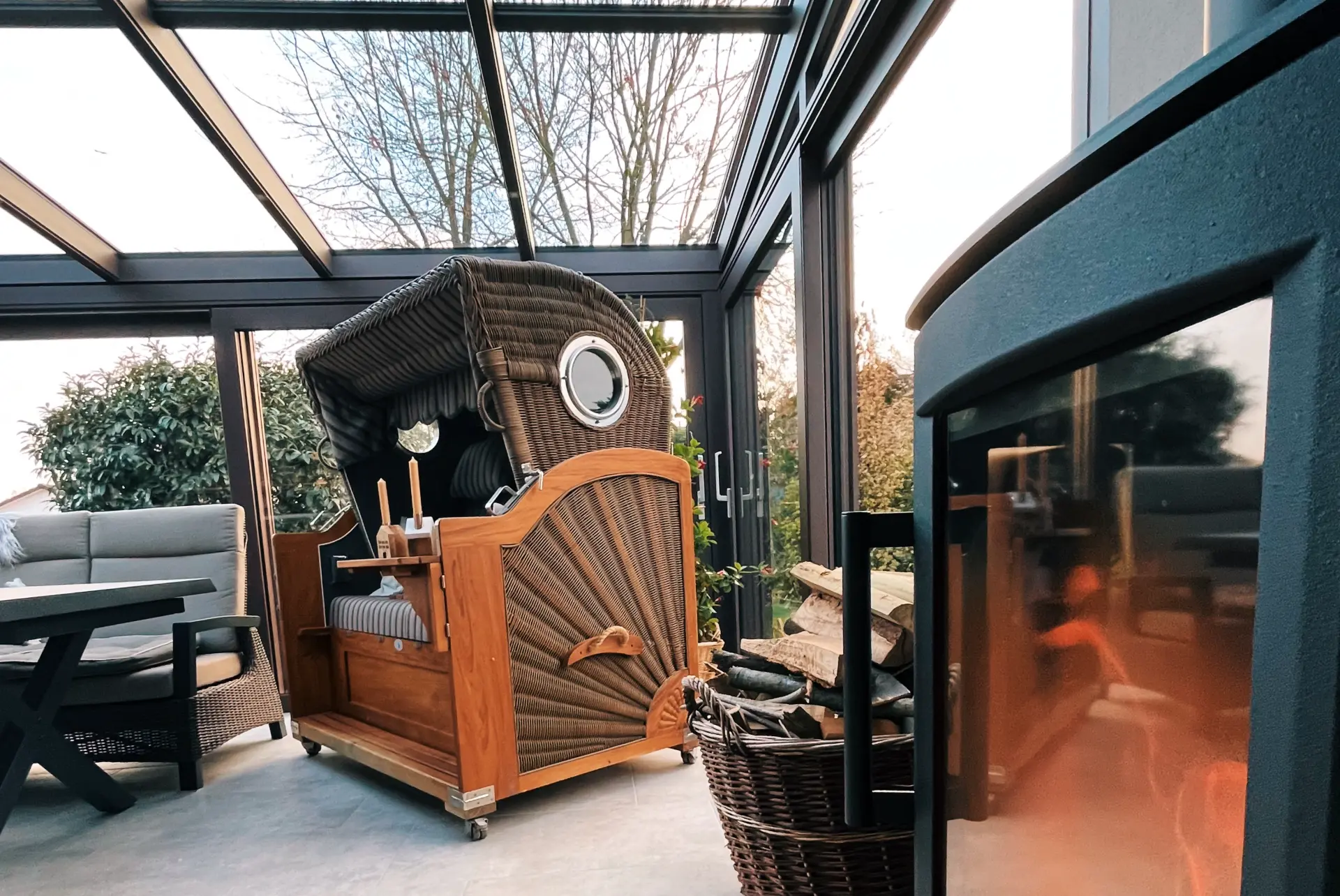 Wintergarten mit Rattan-Strandkorb und Kaminofen unter dem Glasdach, dahinter kahle Bäume im Abendlicht