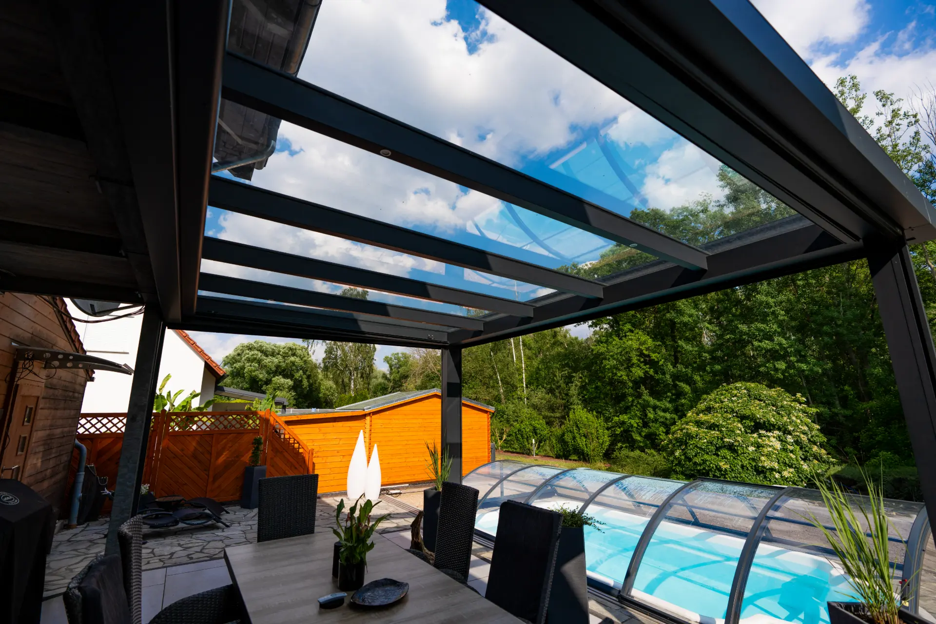 Blick von der Terrasse unter einem Glasdach aus Aluminium auf den Pool und den Garten dahinter
