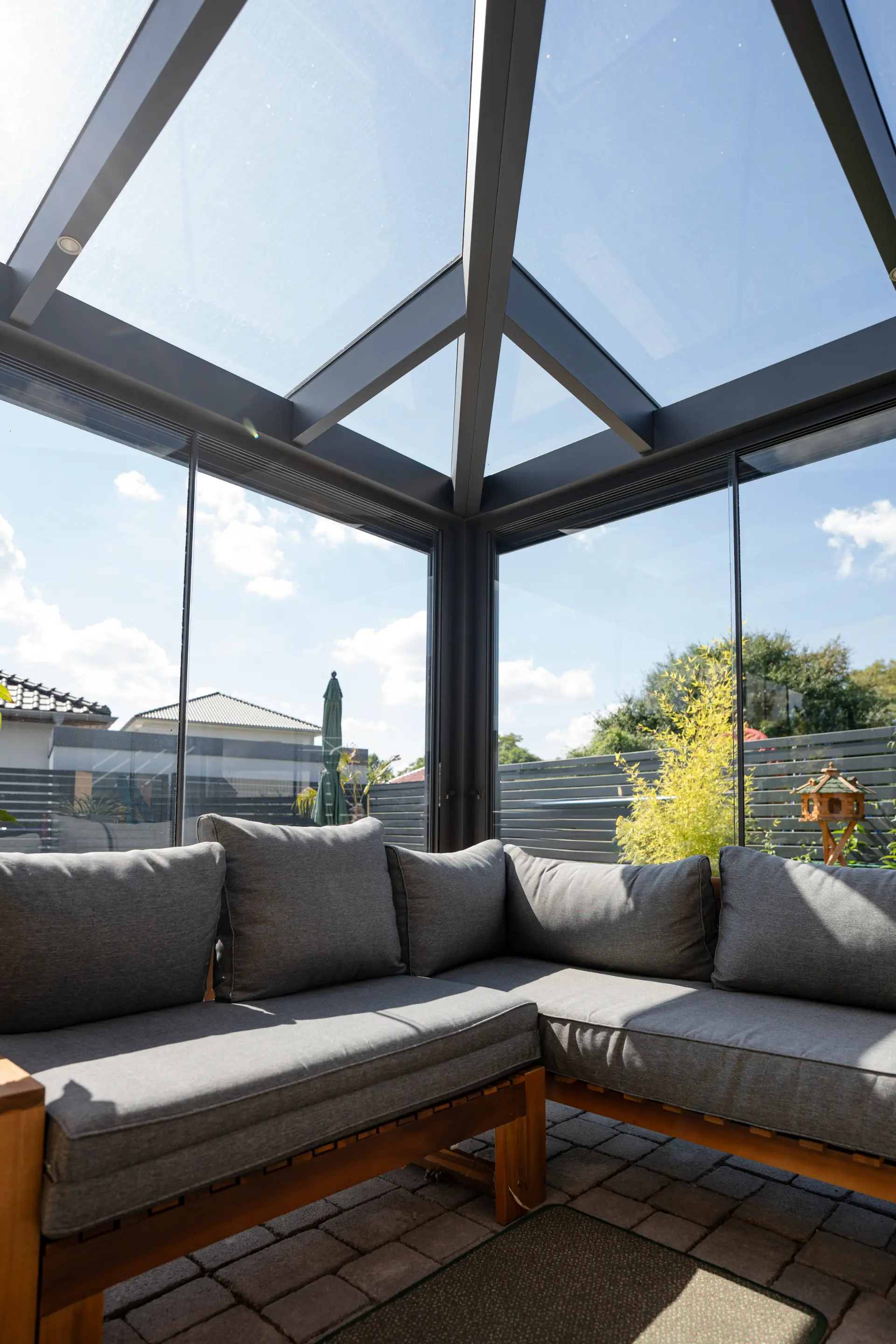 Vollverglaste Wintergartenecke mit grauer Sitzbank, dahinter der sonnige Garten mit Sonnenschirm