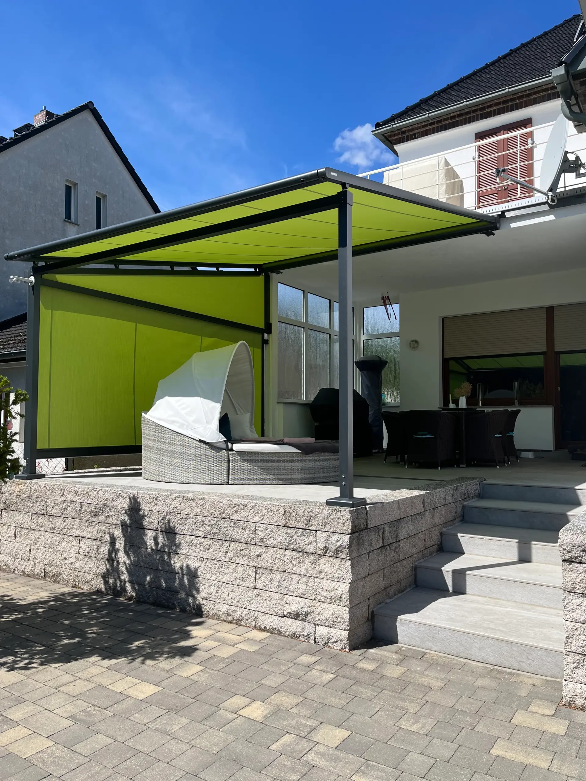 Grüne Pergolamarkise auf zwei Stützen über einer erhöhten Terrasse, seitlich eine Senkrechtmarkise als Sichtschutz
