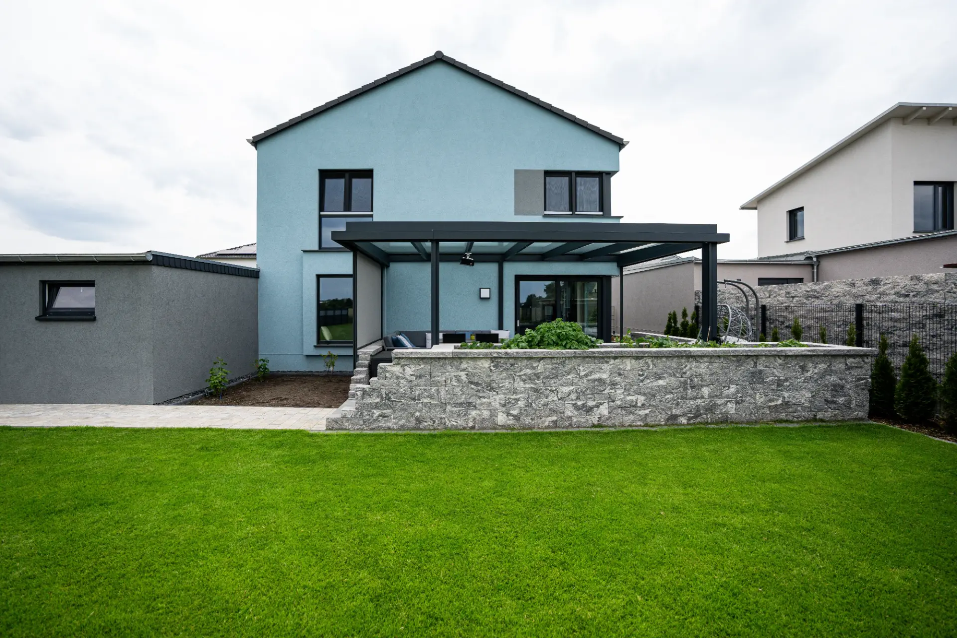 Terrassendach in Anthrazit mit offenen Screens an einem hellblauen Haus, davor eine Natursteinmauer
