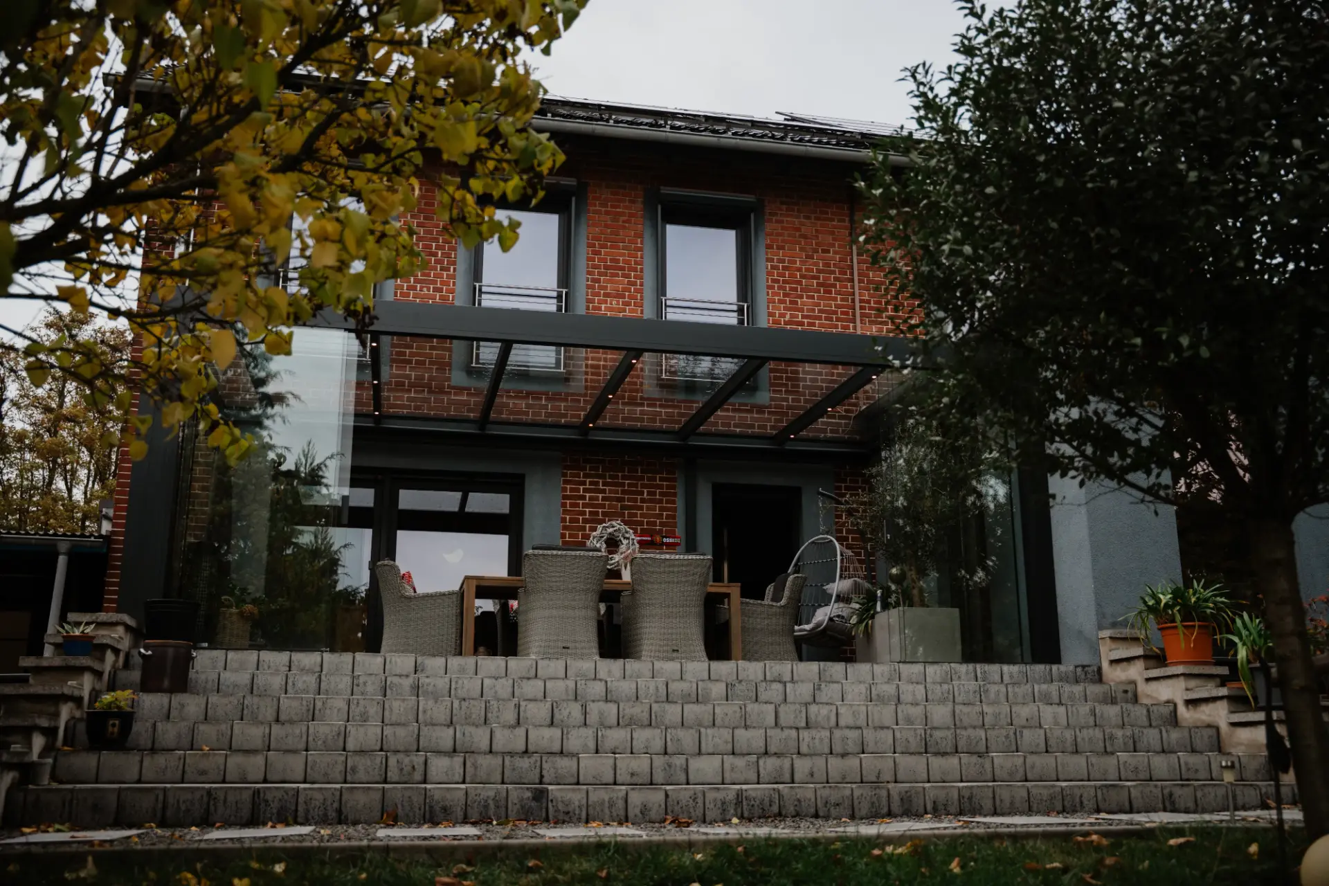Terrassendach aus Glas mit anthrazitfarbenen Profilen an einem Klinkerhaus, darunter eine Rattan-Essgruppe